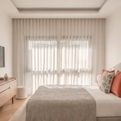 Chambre ensoleillée avec tête de lit cintrée, coussins corail, commode en noyer et rideaux voile sol-plafond