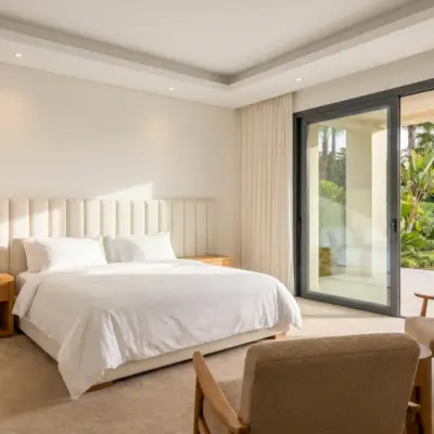 Chambre lumineuse avec tête de lit ivoire, chevets en chêne, fauteuils bouclé et baies vitrées sur jardin