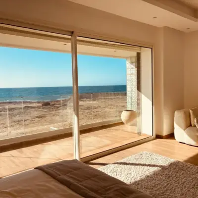 Chambre parentale avec baie toute hauteur ouvrant sur une vue mer panoramique, fauteuil en bouclé crème et tapis en laine