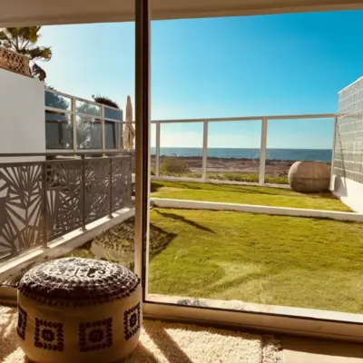 Vue depuis le salon à travers une baie vitrée sur la pelouse clôturée face à la mer, avec pouf brodé et tapis berbère